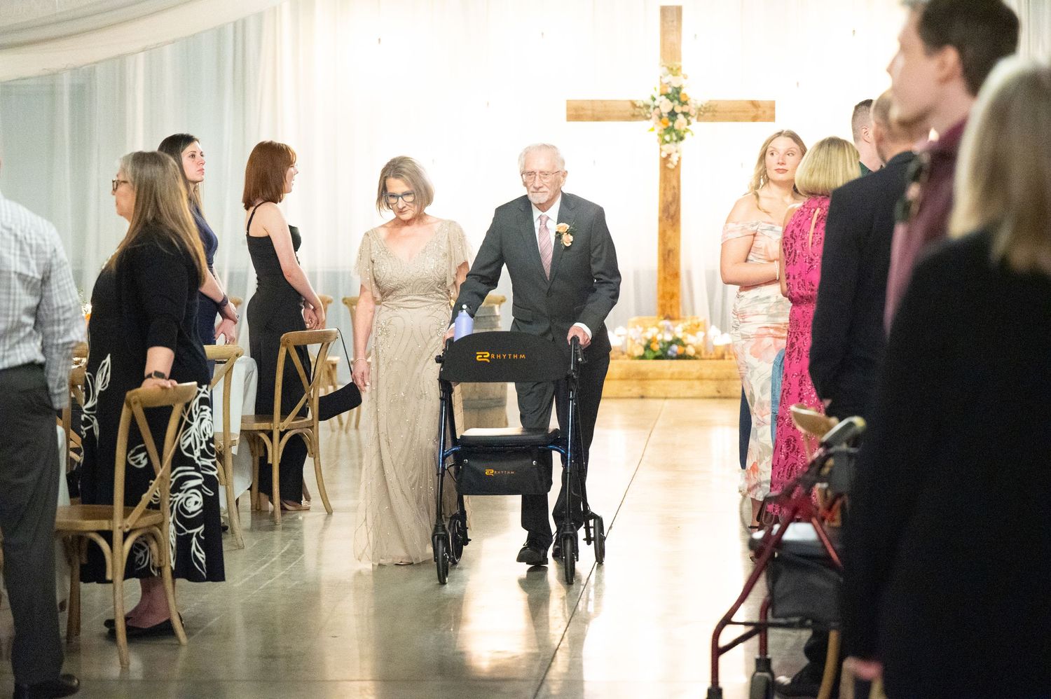 Elderly man with walker escorted by woman in silver gown down wedding ceremony aisle.