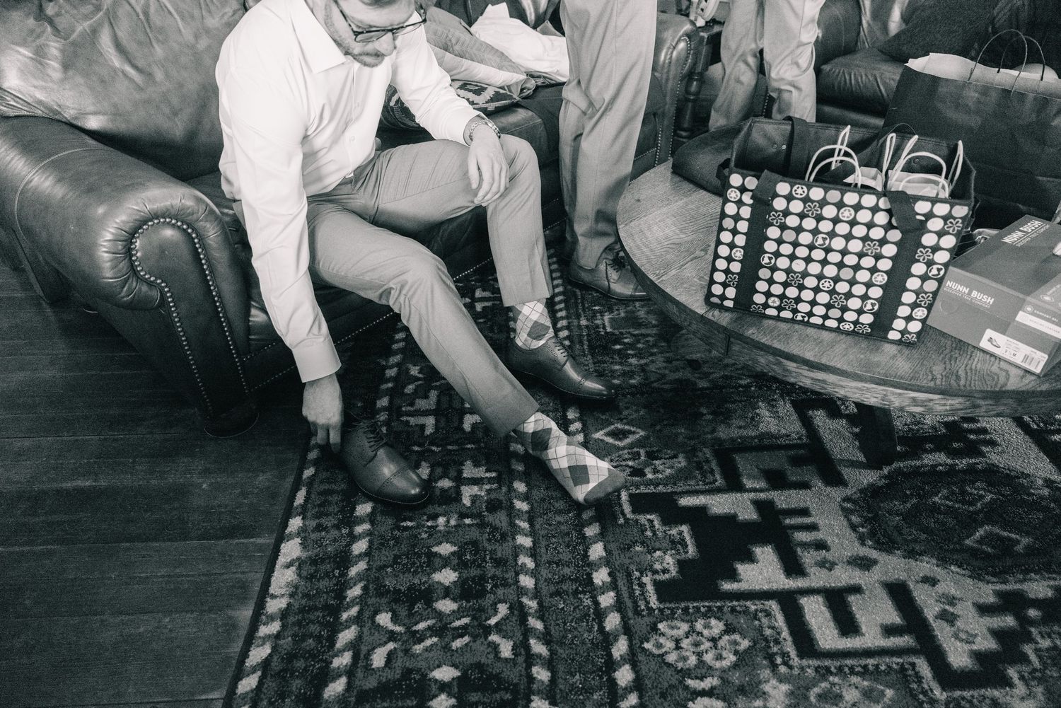 Groom putting on dress shoes while seated on leather couch on patterned rug before wedding ceremony.