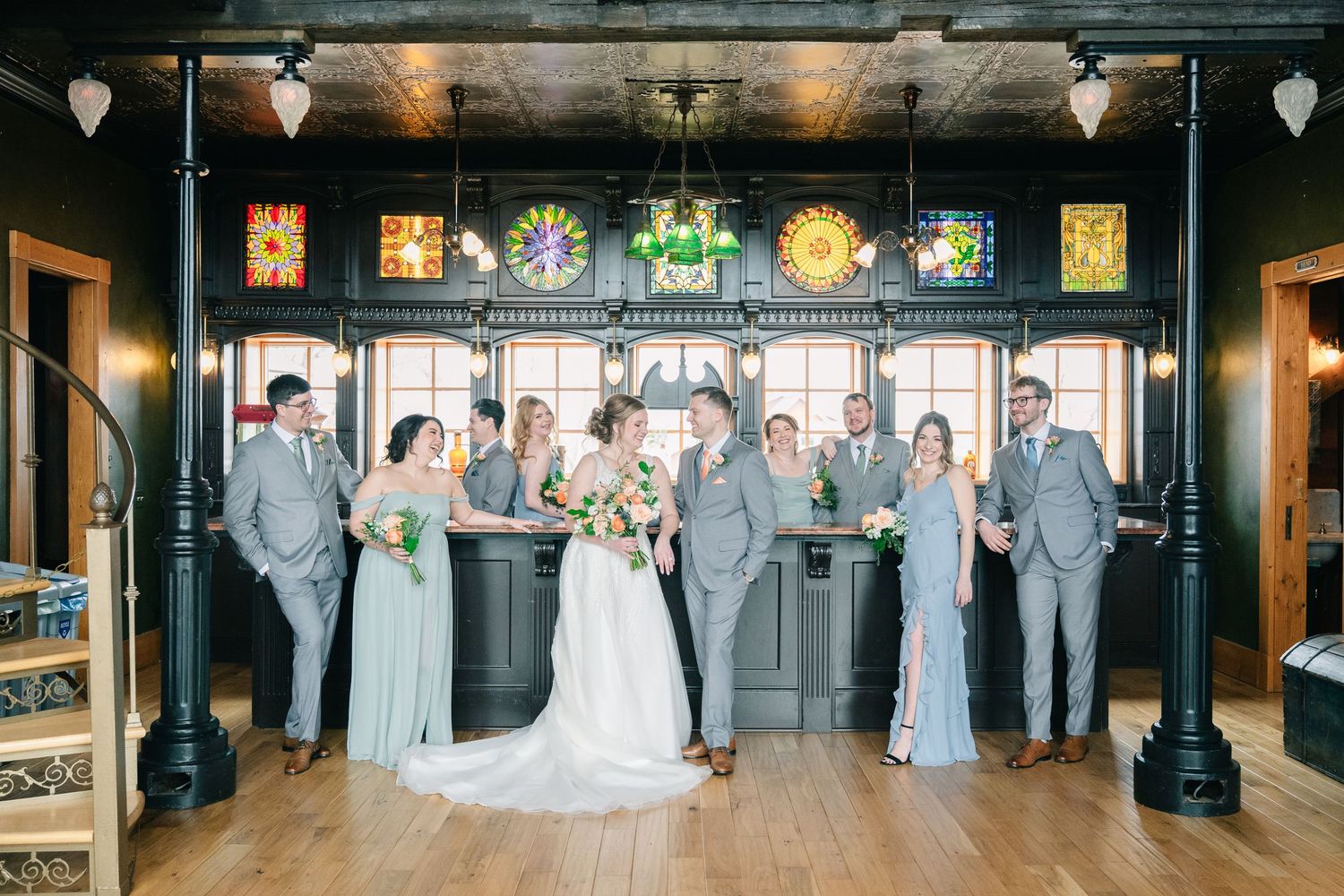 Wedding party in gray attire posing inside a venue with colorful stained glass windows and dark decor.