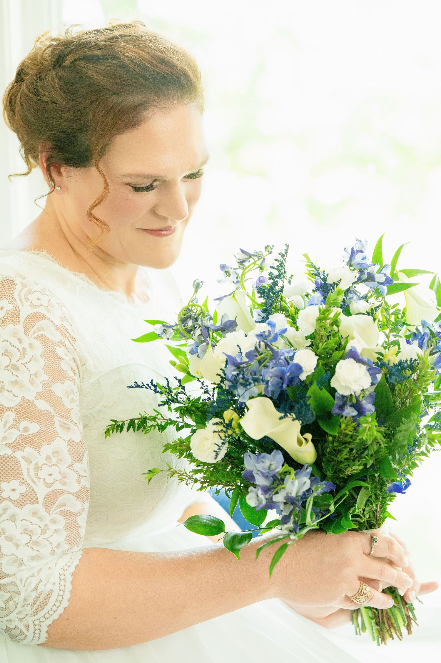 Bride in lace wedding dress holding blue and white floral bouquet with greenery, looking down softly.