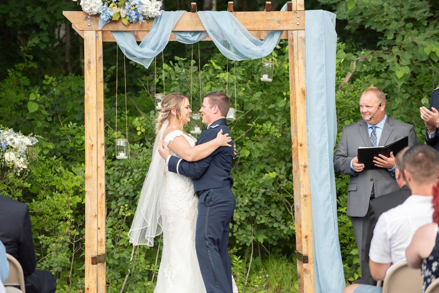Outdoor wedding ceremony under a wooden arch decorated with light blue fabric and floral arrangements in a garden setting. Zimmerman MN Wedd