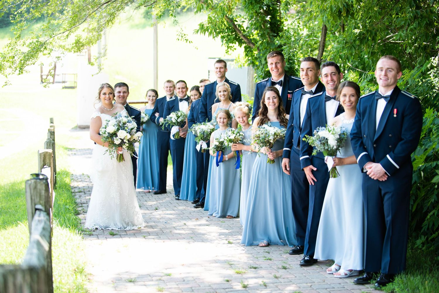 Wedding party in blue dresses and suits lined up outdoors on a garden path. Zimmerman MN Weddings
