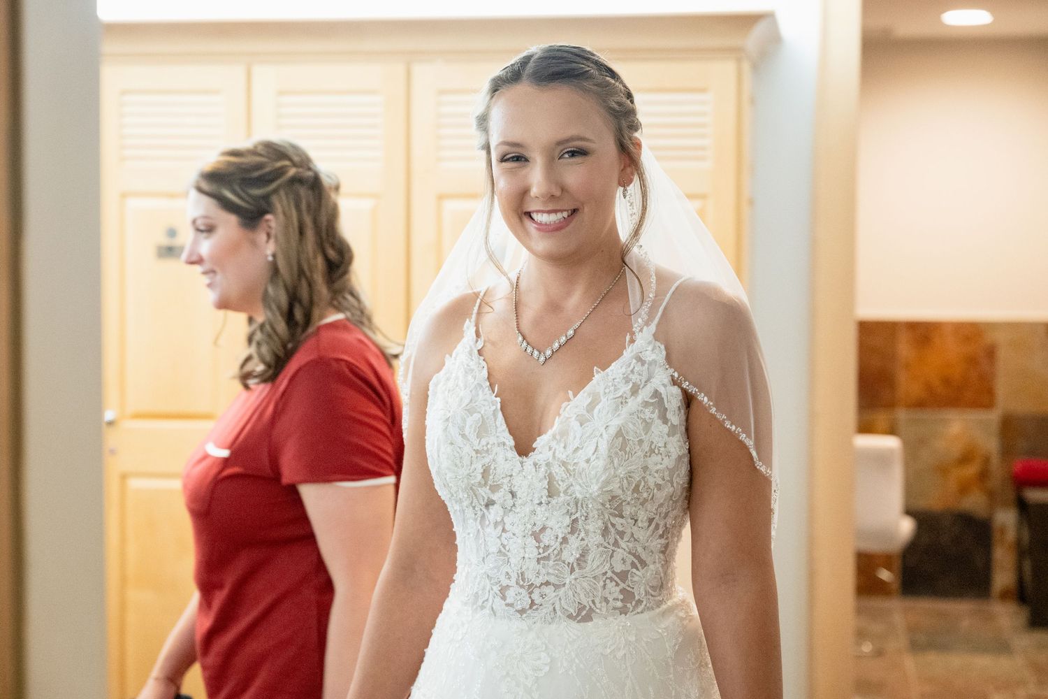 Smiling bride in lace wedding gown with veil, assisted by bridesmaid in red dress.