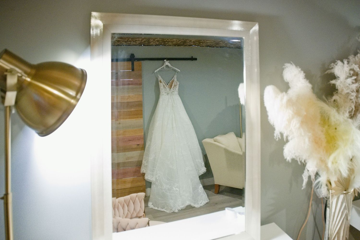 White lace wedding dress hanging on a hanger, reflected in a vanity mirror with gold lamp and pampas grass decor.