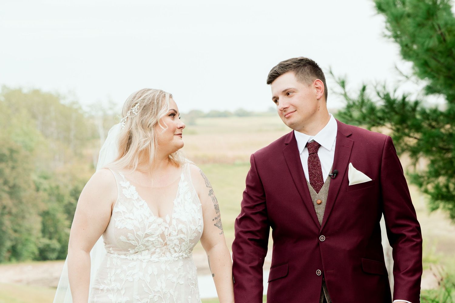 A wedding couple stands affectionately together outdoors in a natural setting with burgundy and cream attire.