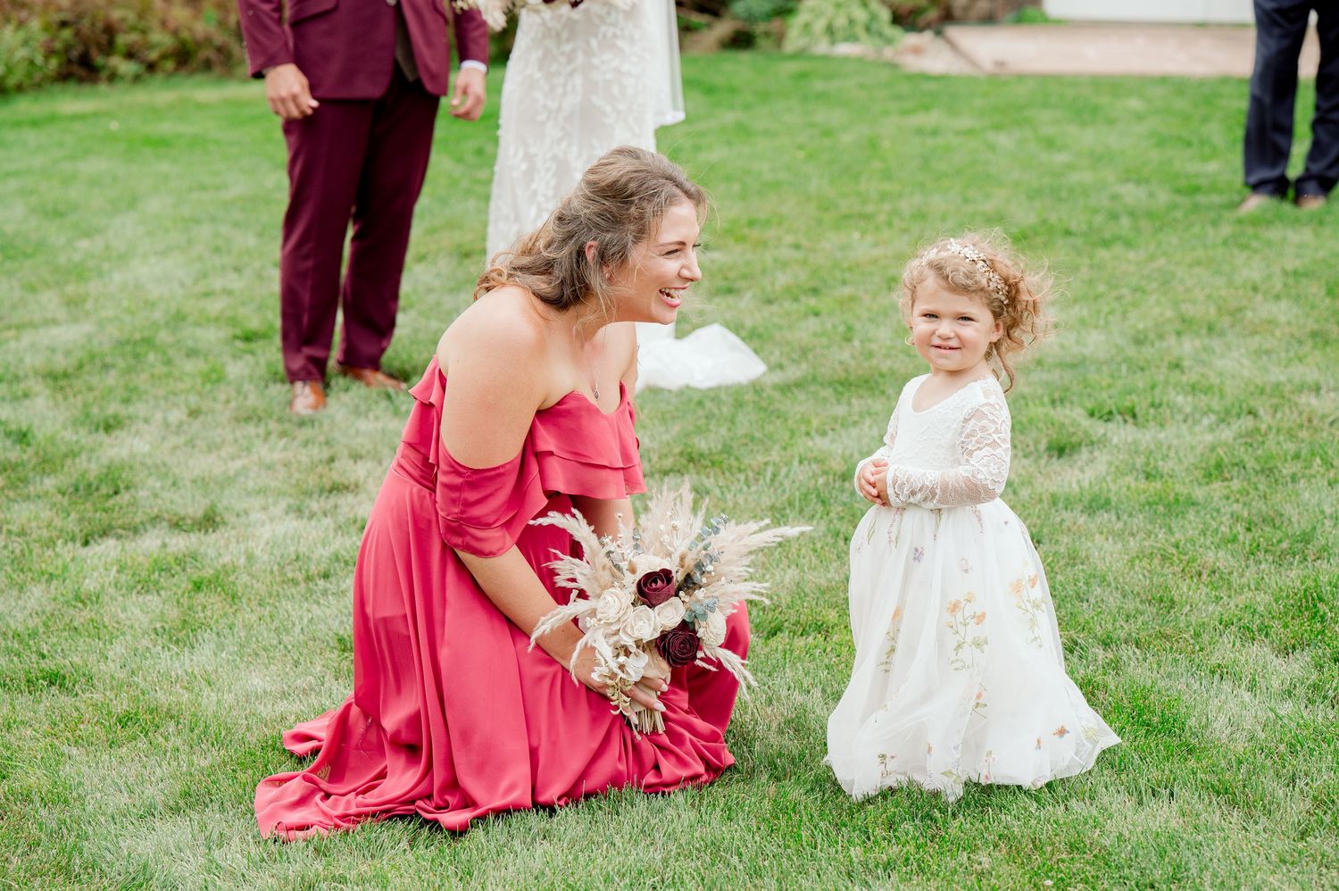 A bridesmaid in a pink dress kneels on grass while interacting with a young flower girl in white.