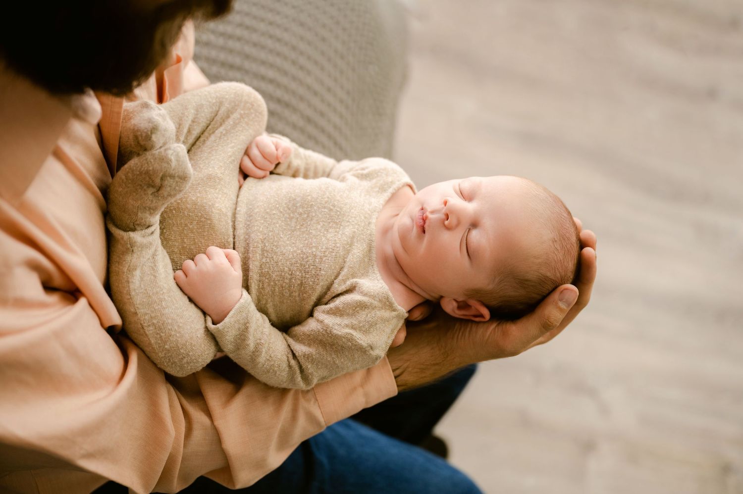 A sleeping baby wearing beige clothing rests peacefully in arms while being cradled.