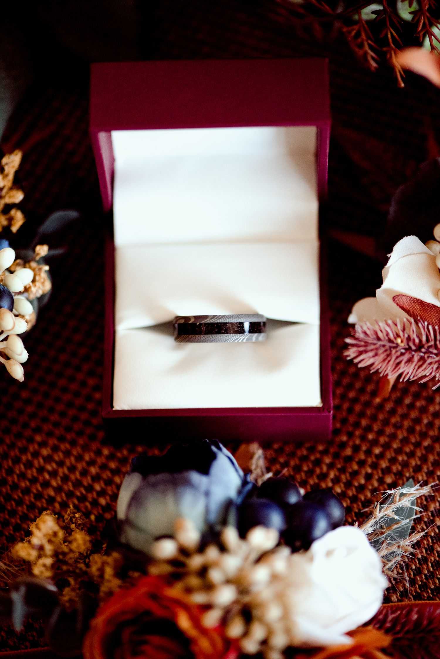 Wedding ring nestled in a burgundy ring box surrounded by seasonal florals and decor on a textured surface.