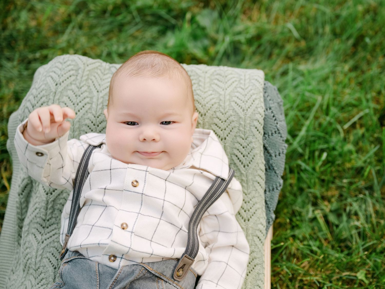 Adorable baby boy in suspenders lying on a knit blanket outdoors on green grass, smiling and reaching up.. Minnesota milestone photographer