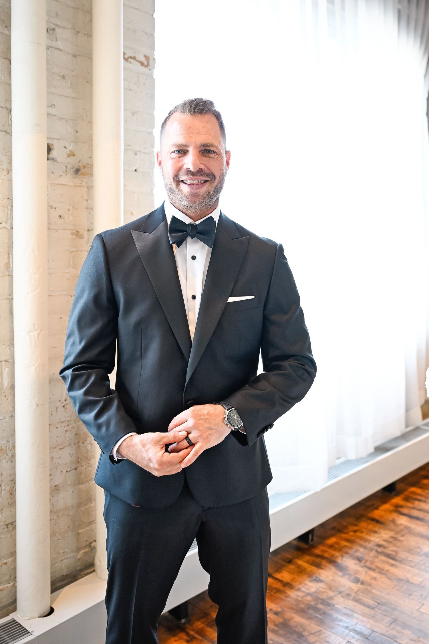 Groom in black tuxedo smiles while standing between white columns in an industrial venue.