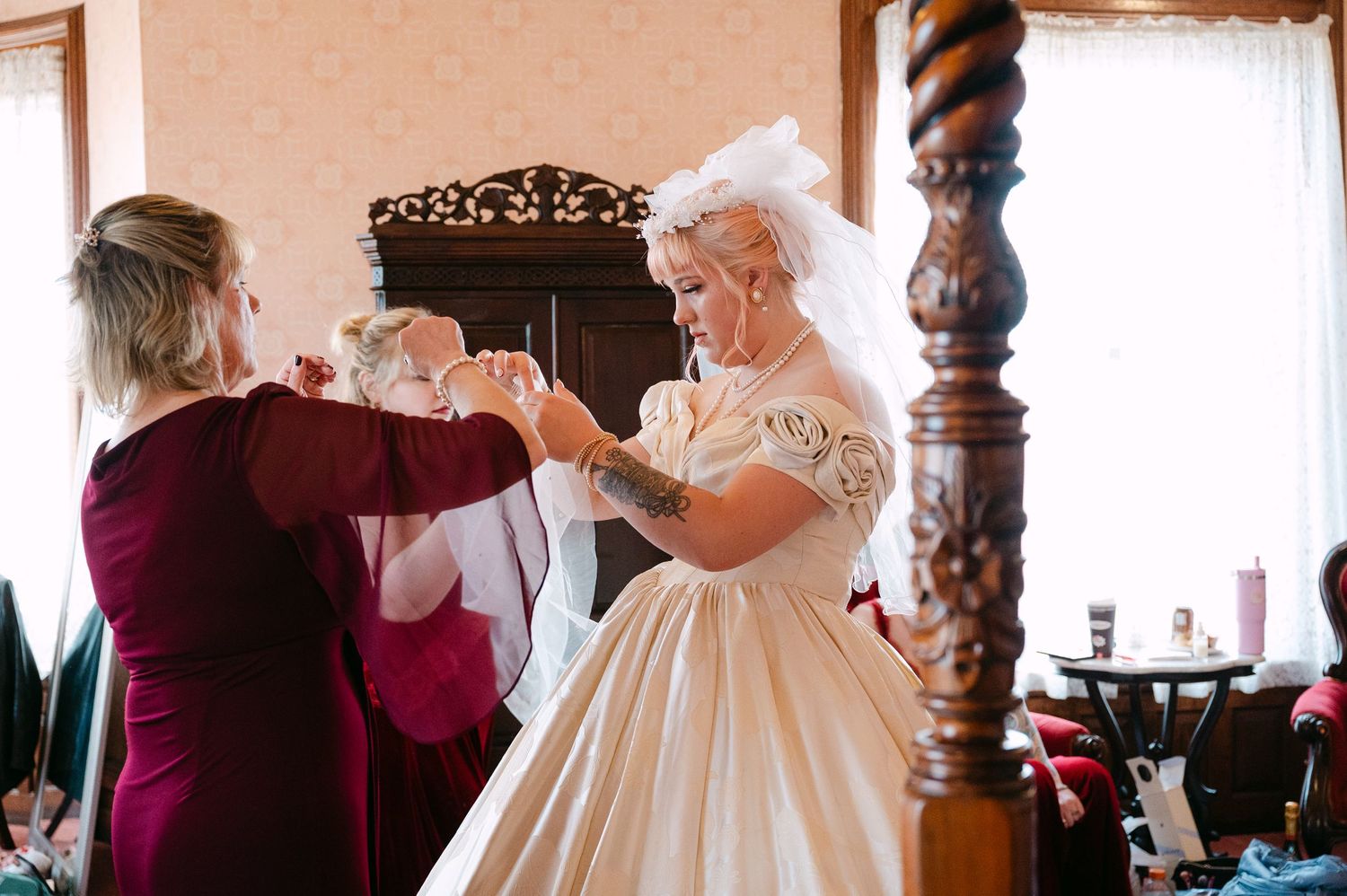 Central MN Wedding Photos | Bride getting help with her gloves from a woman in burgundy dress during wedding preparations in a vintage room.