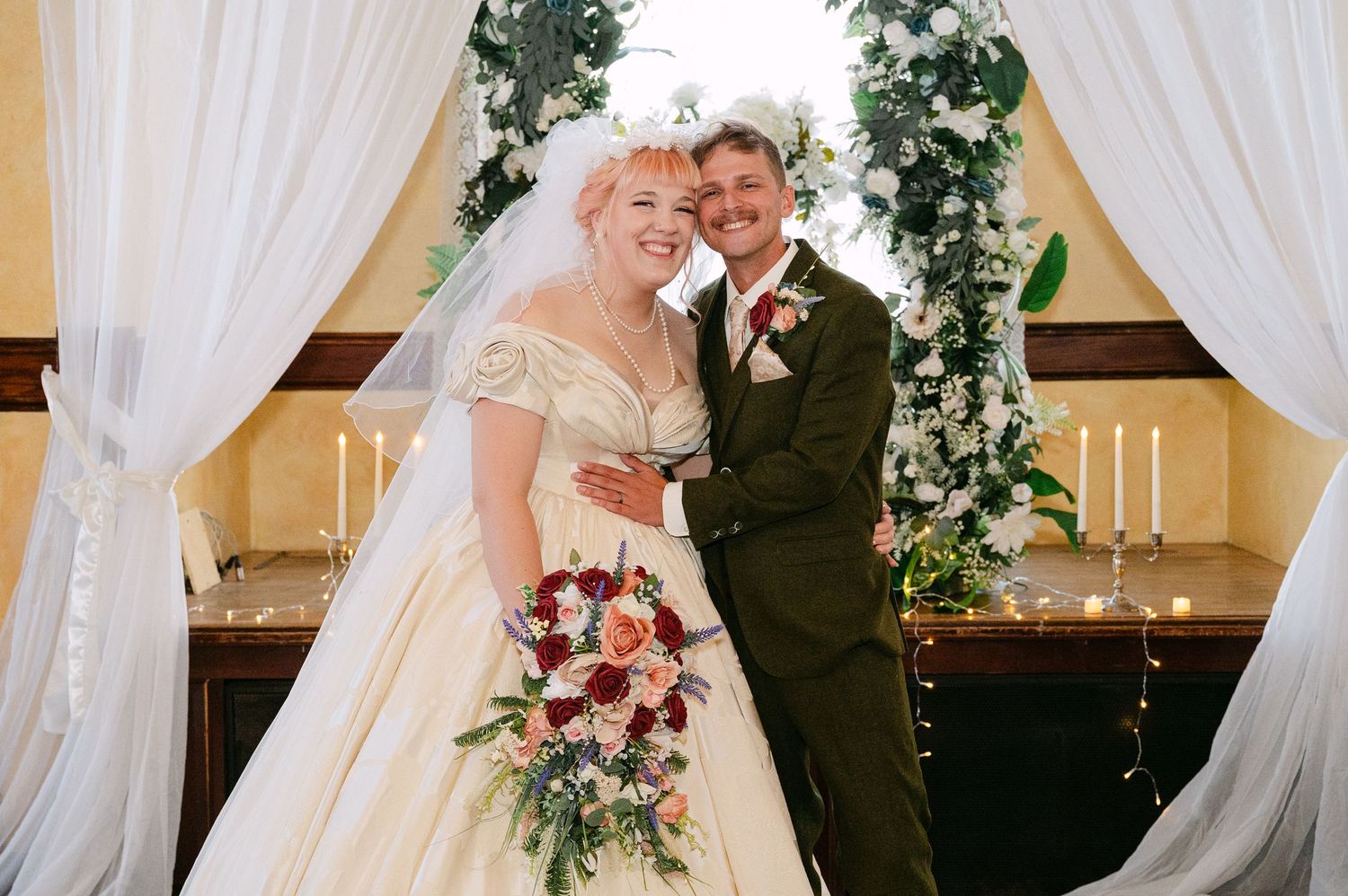Central MN Wedding Photos | Smiling bride and groom pose under floral arch holding colorful wedding bouquet.