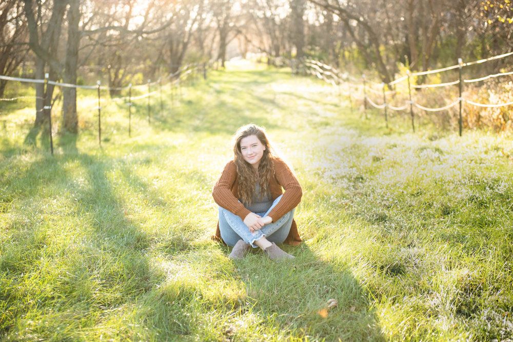 Central MN Senior Photography - Young woman sitting cross-legged on green grass in a sunlit field, wearing a brown jacket and jeans.