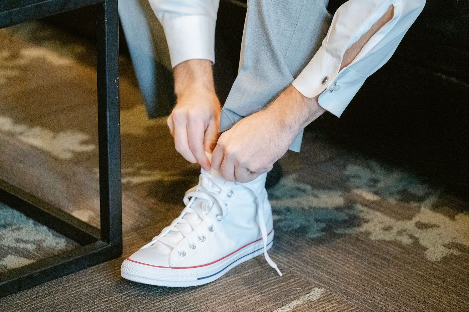 Central MN Wedding Photos | Groom tying white Converse high-top sneakers while wearing a light gray wedding suit.