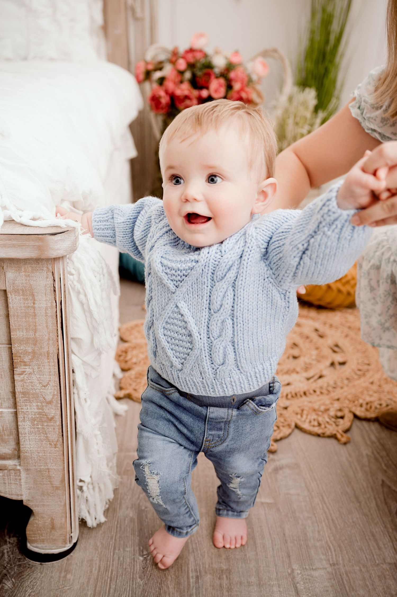 Central MN Milestone Photography - A happy baby in a blue knit sweater and jeans takes first steps in a cozy decorated room.