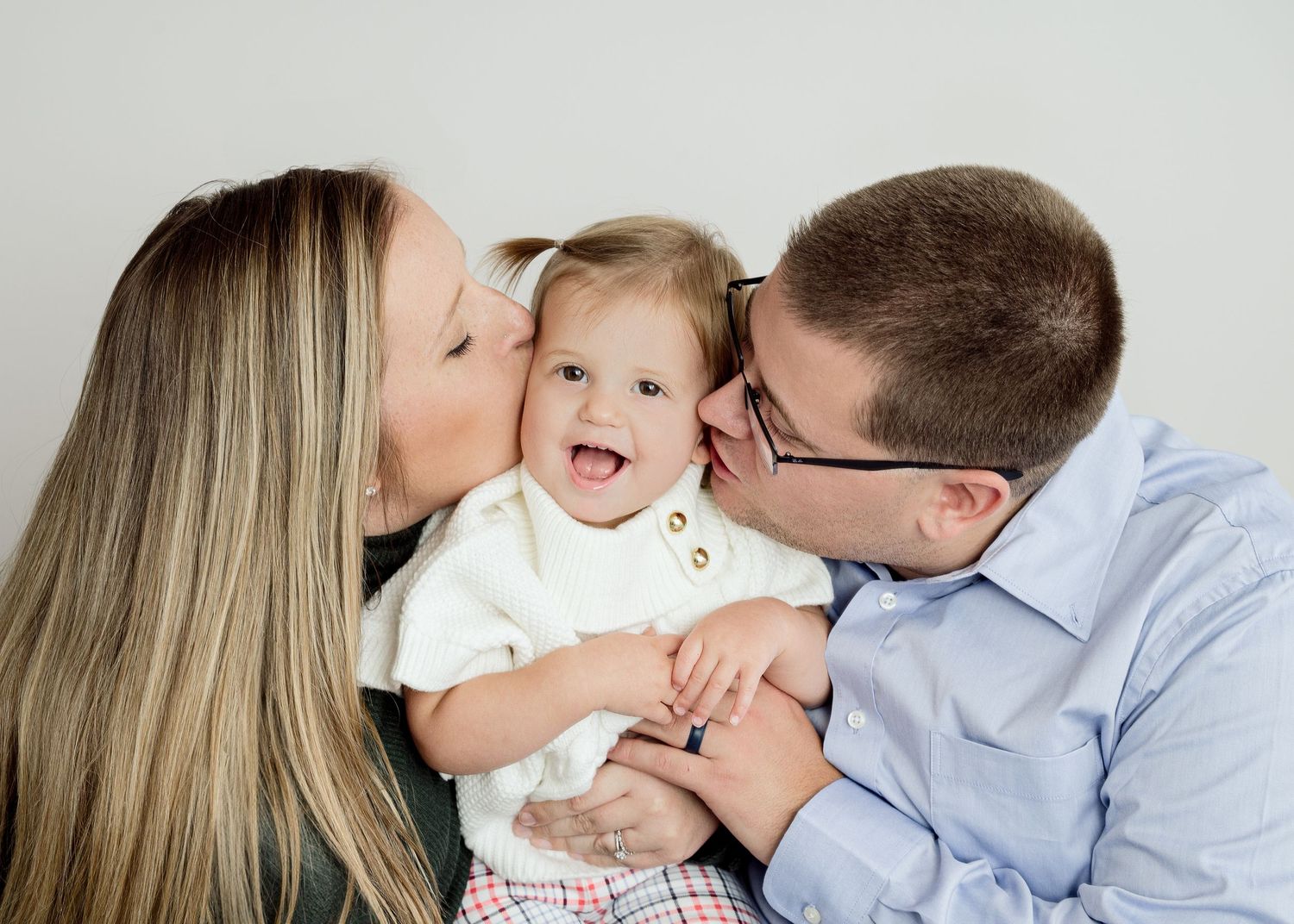 A young child in a white shirt laughs while receiving loving kisses from parents against a light gray background.