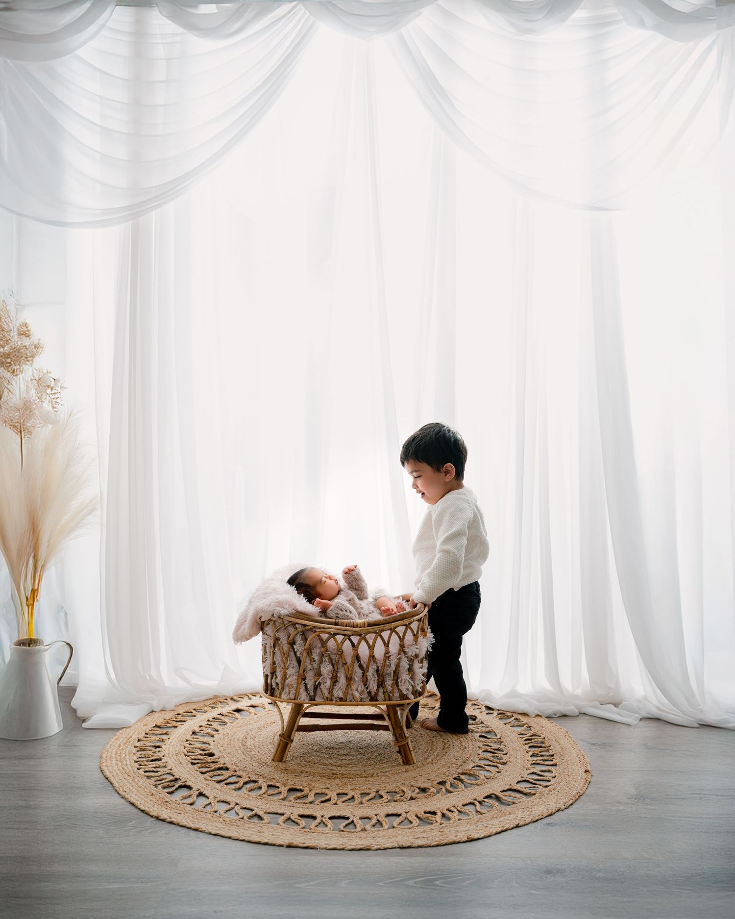 Minnesota Best newborn photographer - Small child looking over a woven bassinet on a decorative round rug against white curtained backdrop.