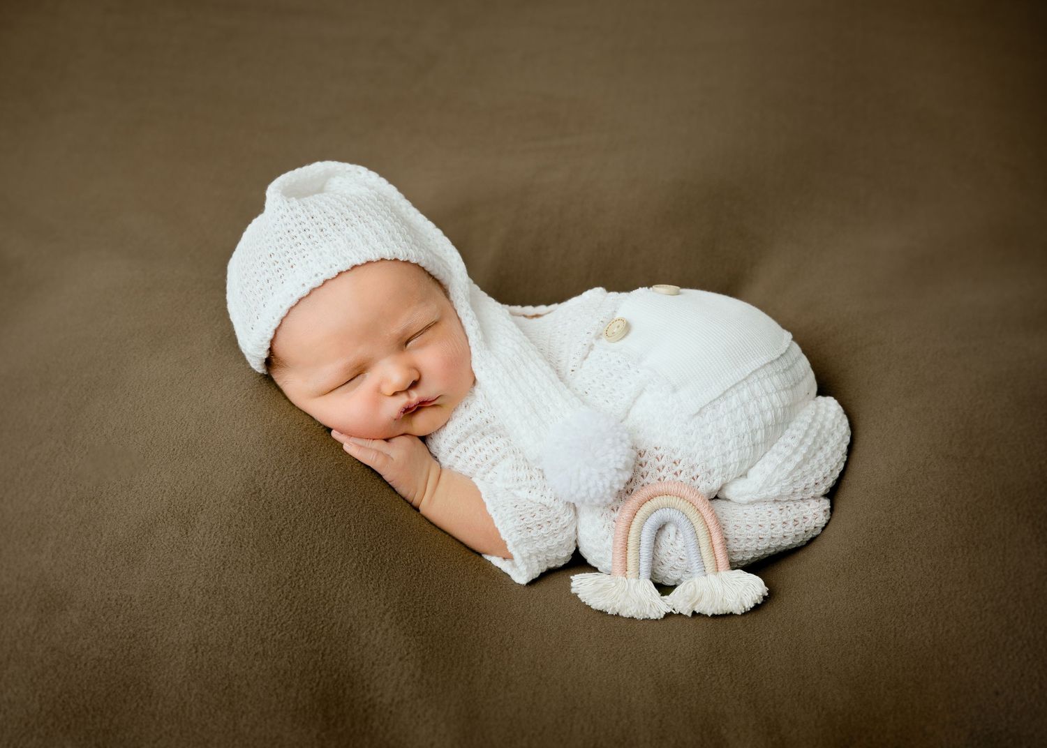 Minnesota Best newborn: sleeping newborn wearing a white knitted outfit and hat lies curled up on a brown backdrop with a small rainbow toy.