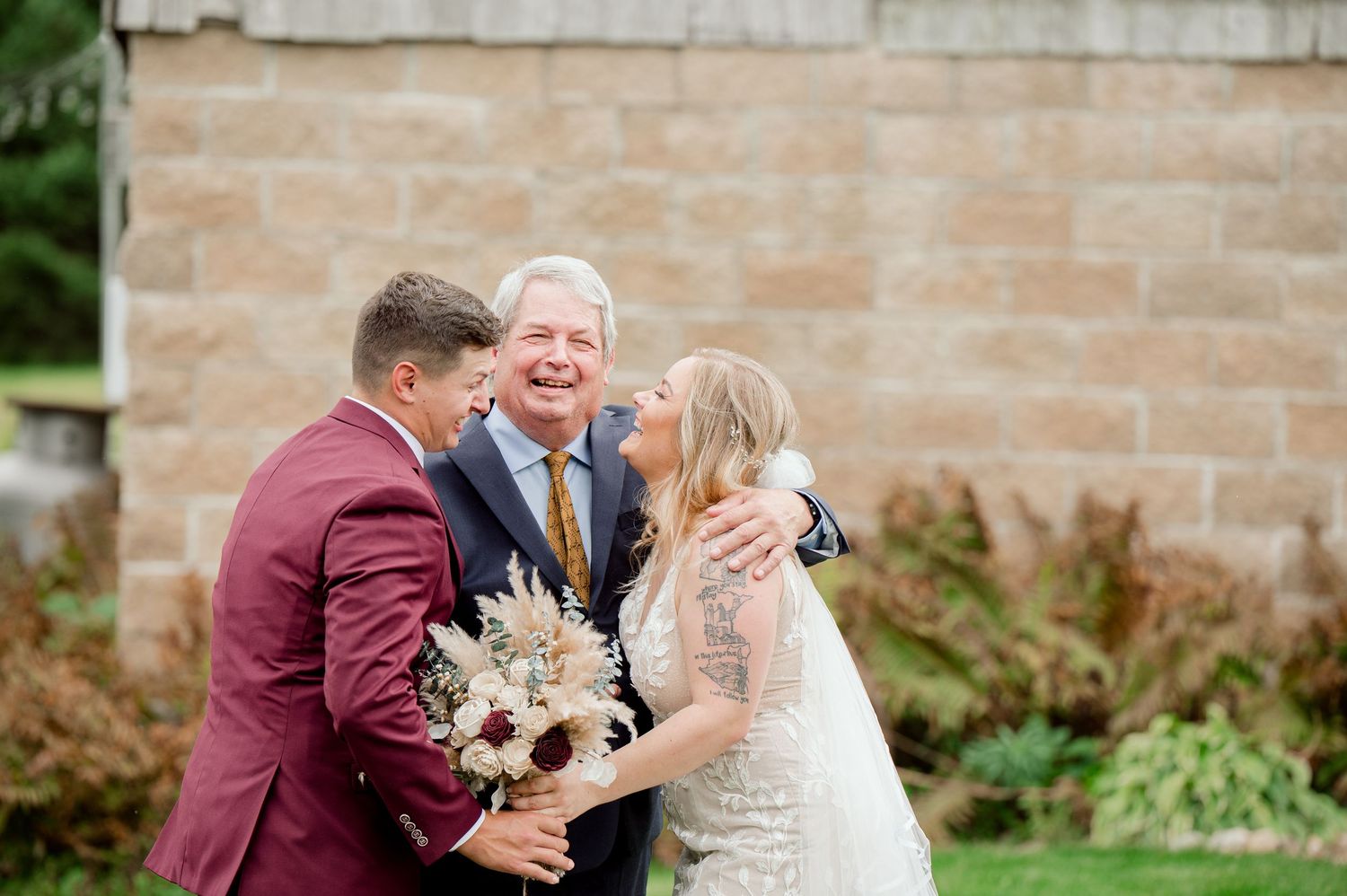 A joyful wedding celebration outdoors with laughter and a beautiful bouquet of dried flowers against a stone wall.