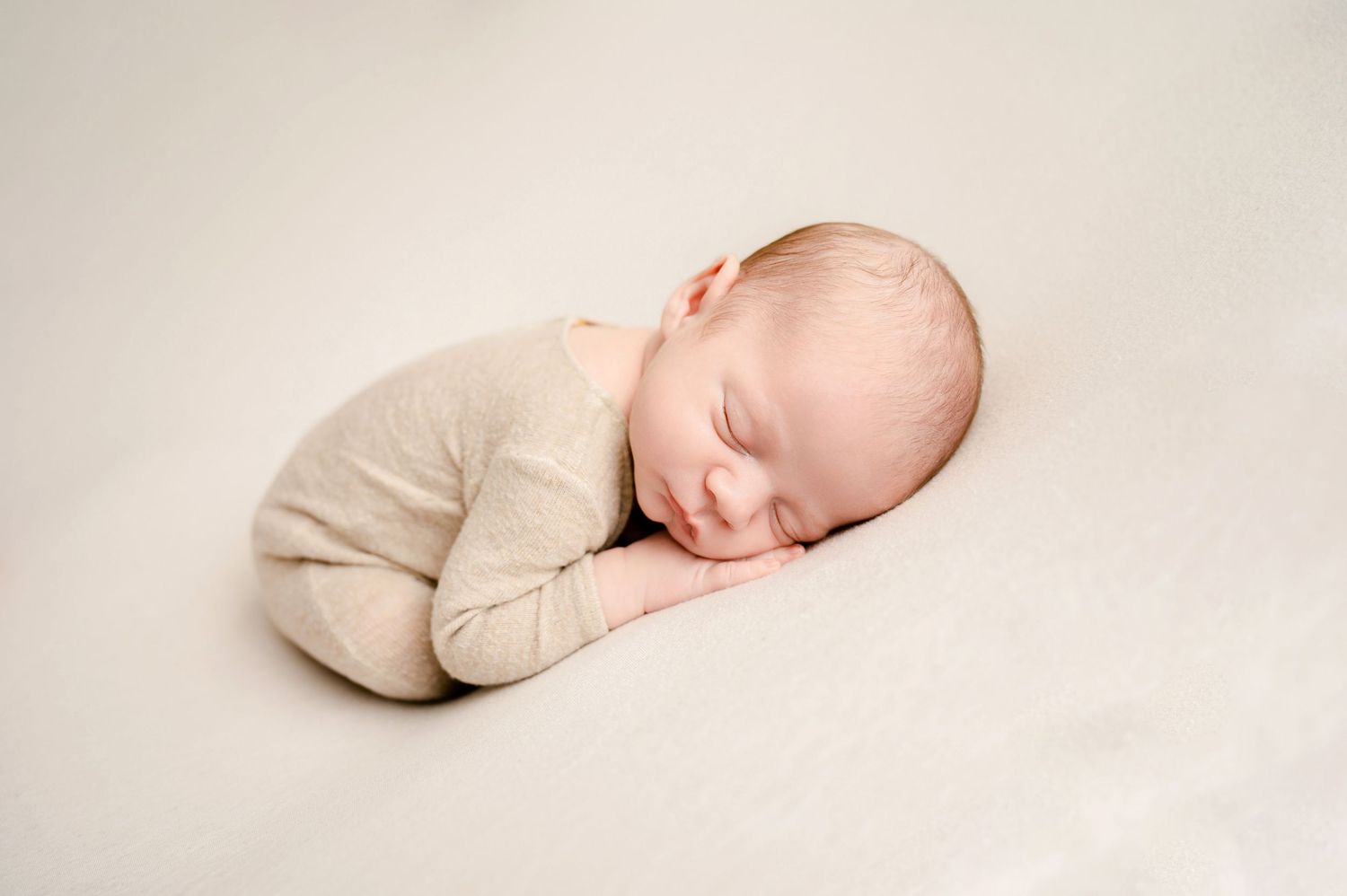 A sleeping newborn baby curled up in a fetal position wearing a cream-colored onesie on light beige fabric.