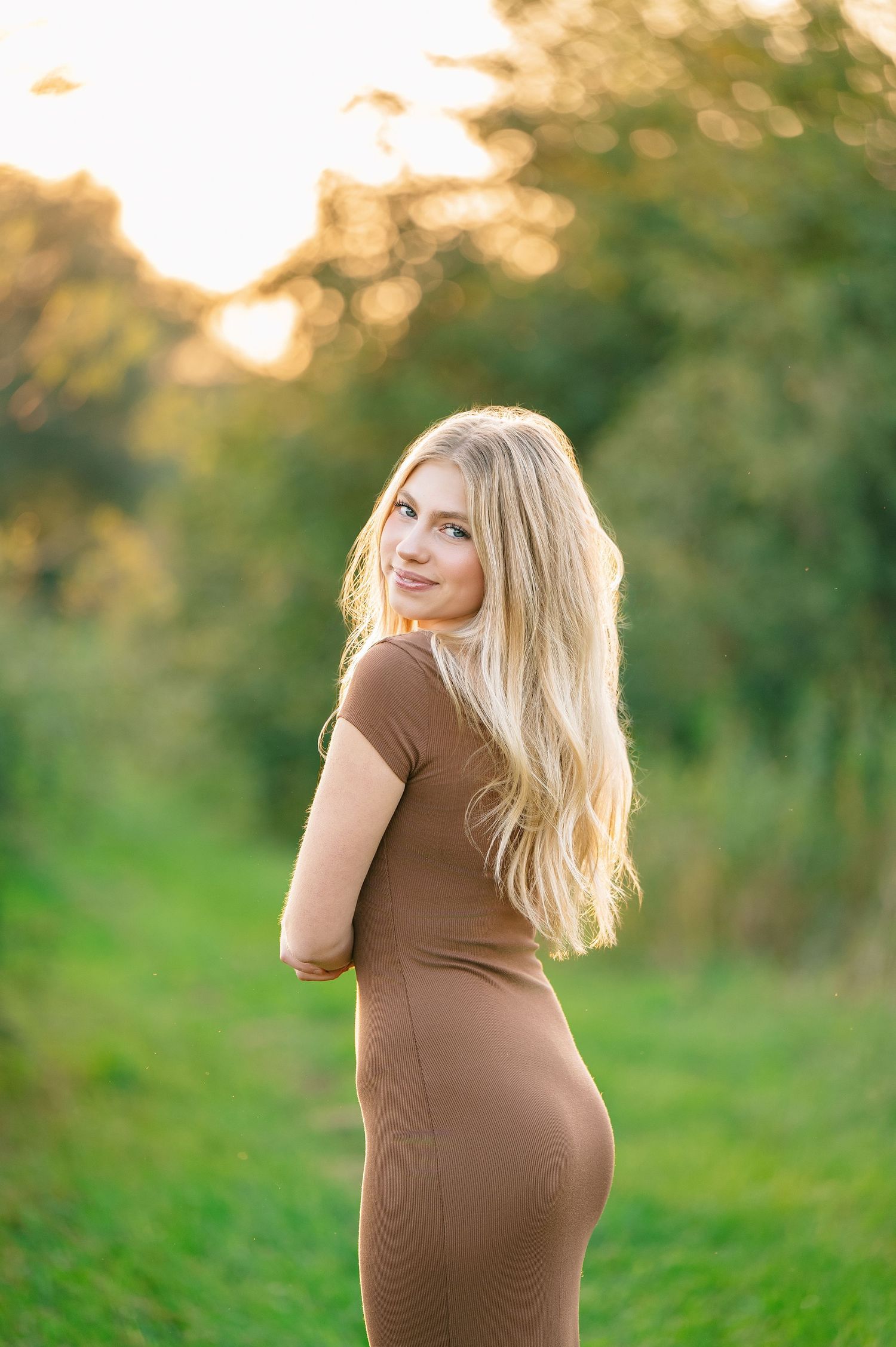 Central MN Senior Photography - Blonde woman in fitted brown ribbed dress standing in sunny green field, smiling over shoulder.