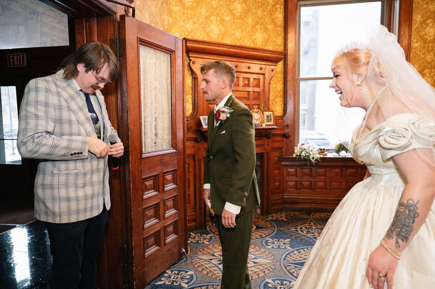 Central MN Wedding Photos | Newlywed couple greeting a guest in plaid jacket inside a historic wooden-paneled venue.