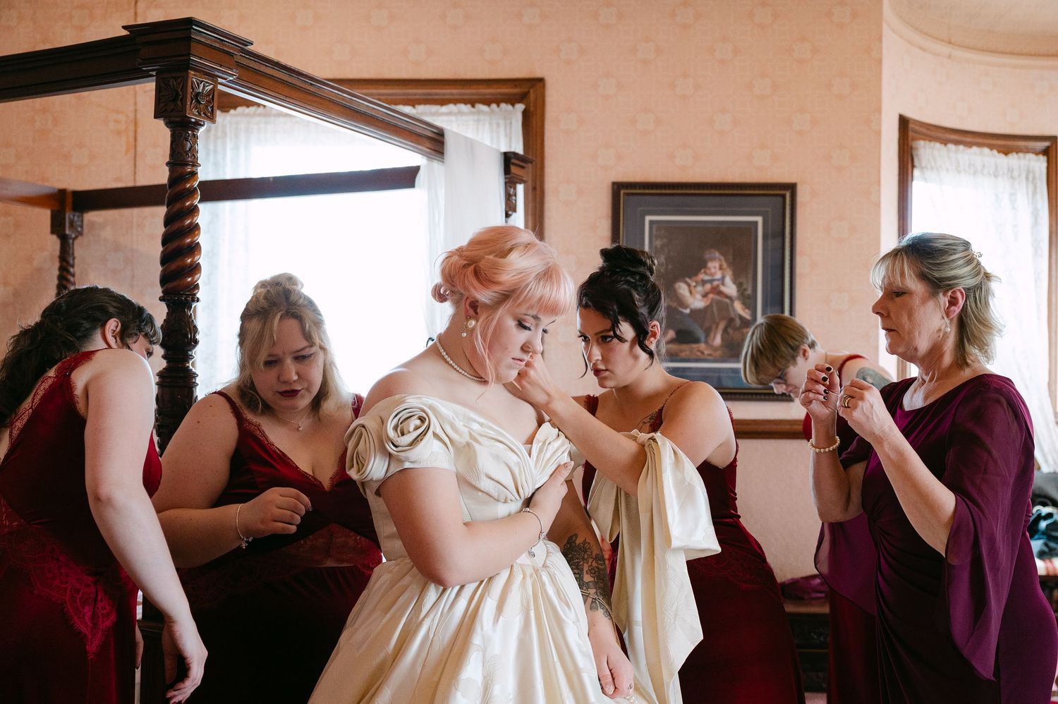 Central MN Wedding Photos | Bridesmaids in burgundy help bride into gold wedding gown while getting ready in elegant room.