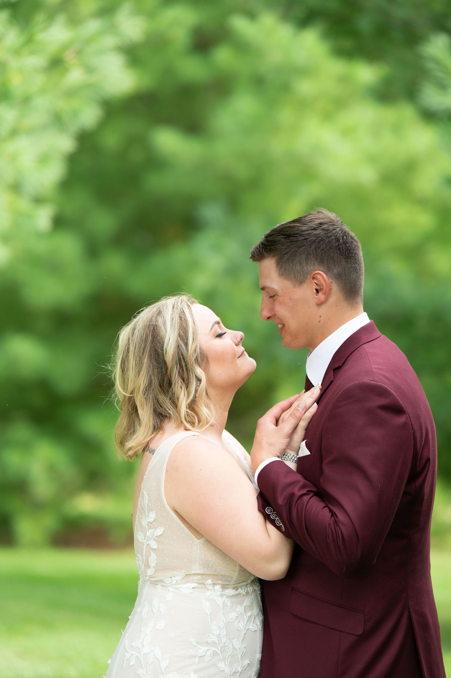 Central MN Wedding Photography - A bride in a white lace gown laughs joyfully with a groom in a burgundy suit outdoors.