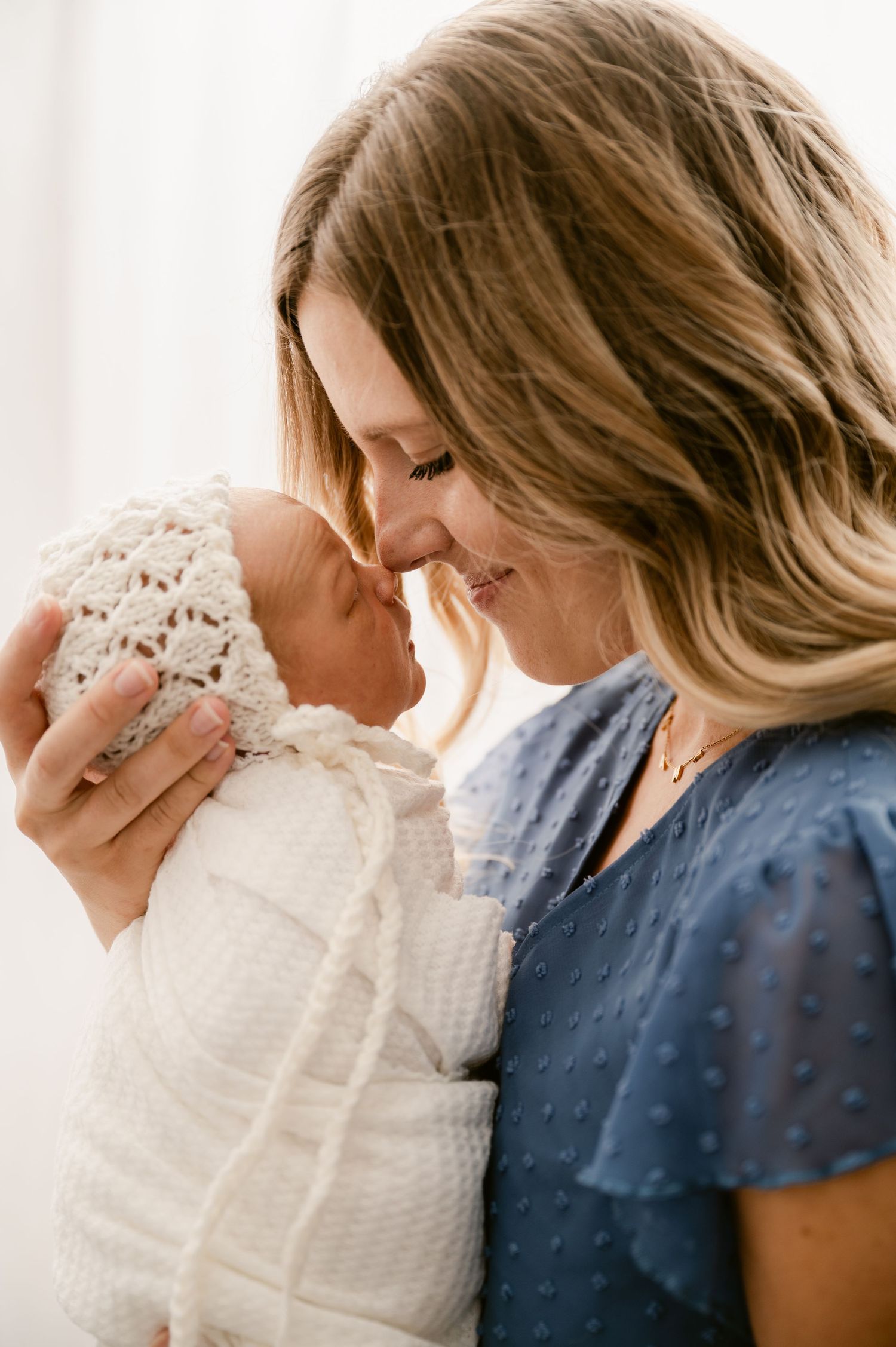 A smiling mother in a blue top tenderly holds her swaddled newborn baby wrapped in a white crochet blanket.