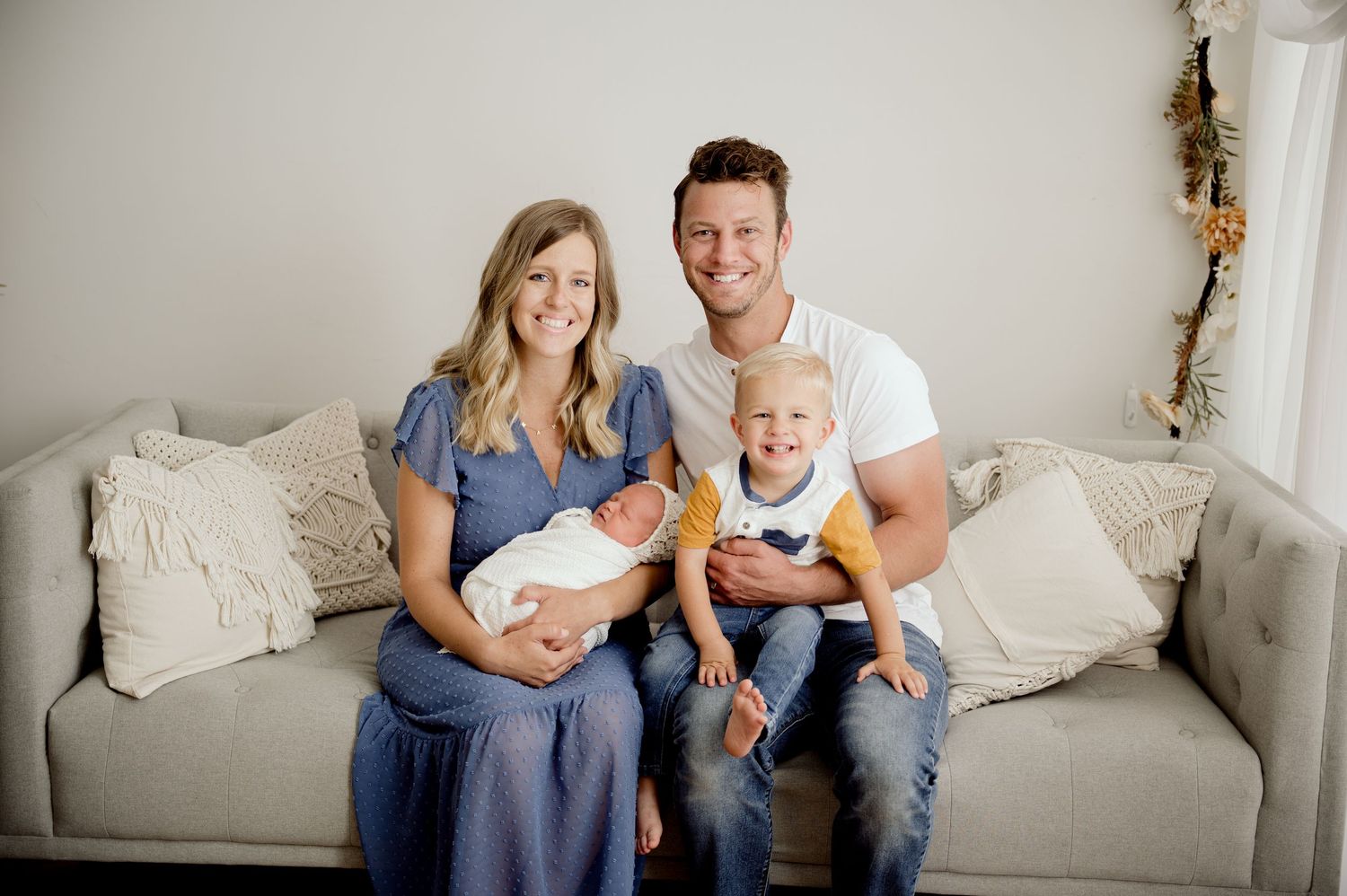 Family of four posing on a couch, mom holding newborn baby, dad holding toddler, smiling in a bright studio setting.