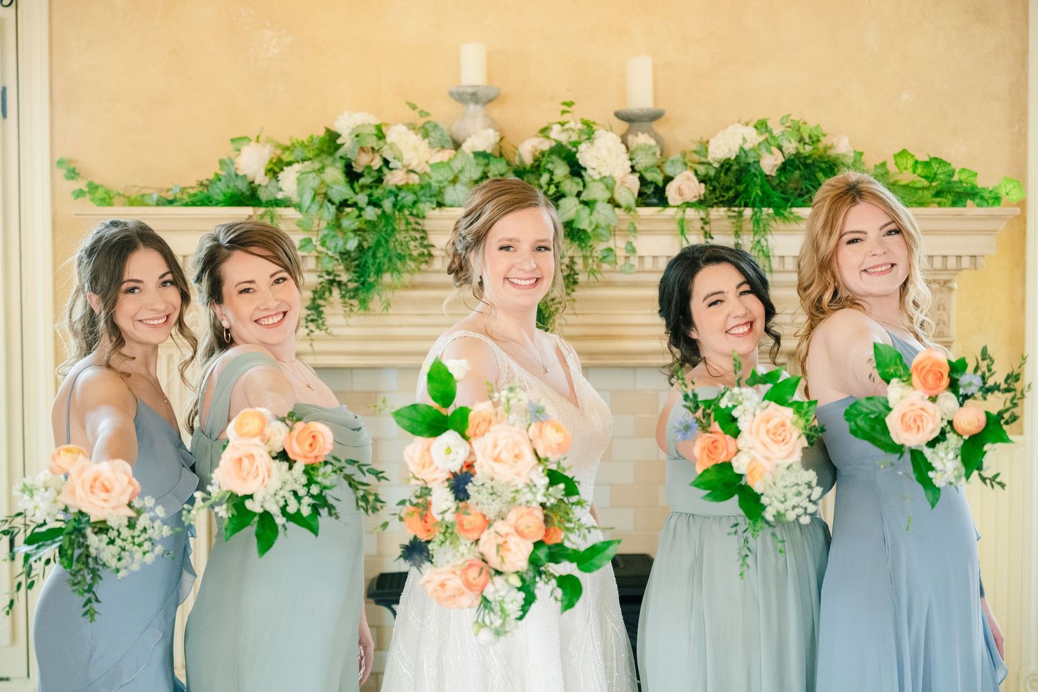 Bride with four bridesmaids in dusty blue gowns holding peach bouquets before floral fireplace.