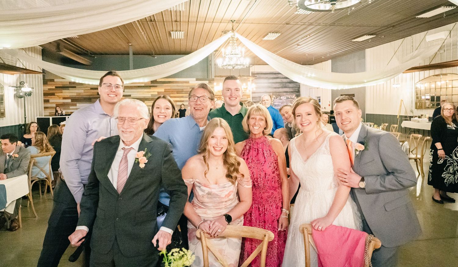 Wedding party and family gather for a group photo in a decorated reception hall.