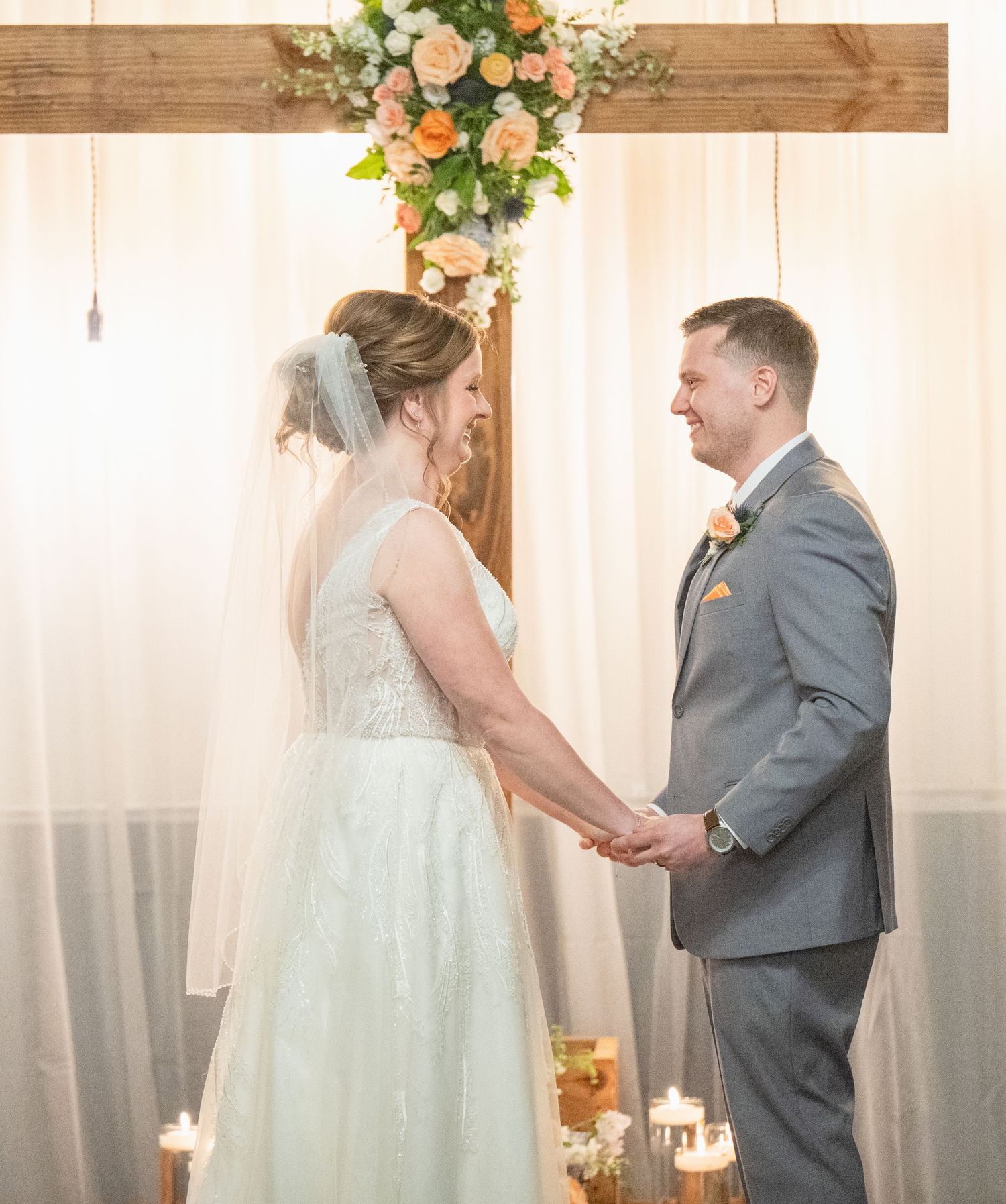 Bride and groom holding hands at the altar beneath a wooden arch with peach floral arrangements and candles.