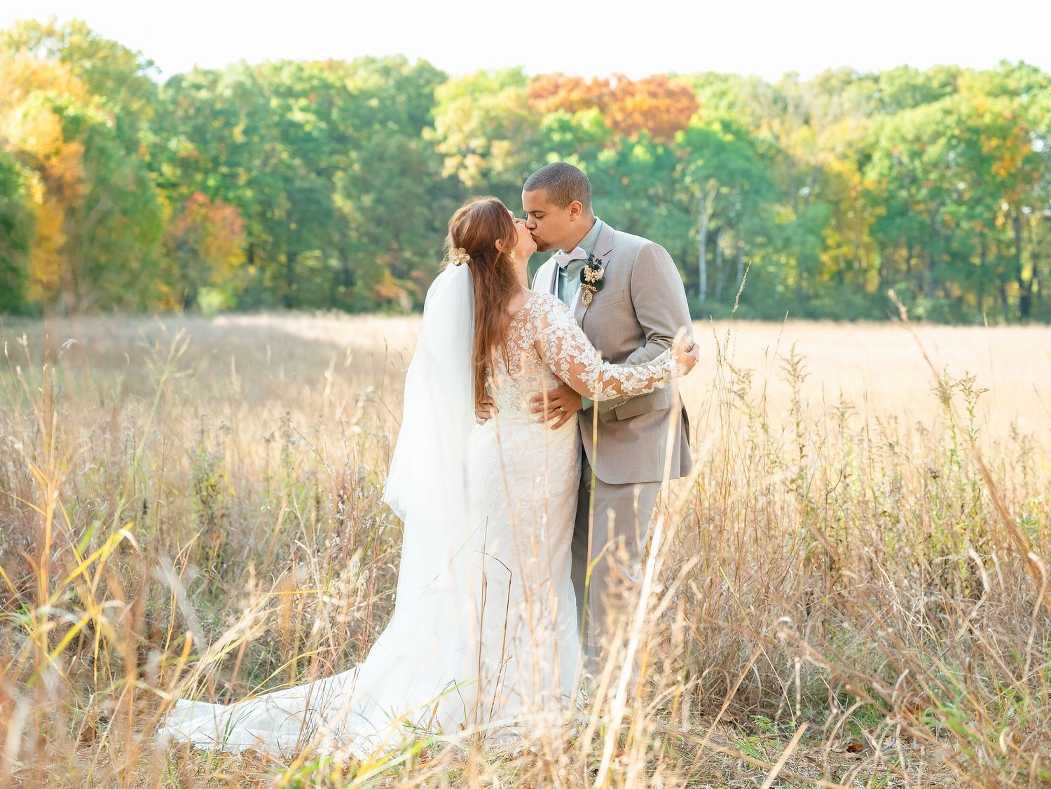 Central MN Wedding Photos | A bride and groom share a romantic kiss in a golden autumn field with colorful fall foliage in the background.