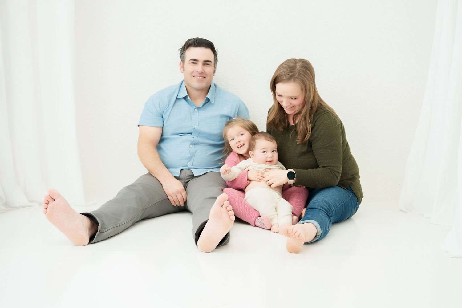 Happy family of four sitting on white floor, parents smiling with two young children in bright studio photo.. Minnesota milestone photograph