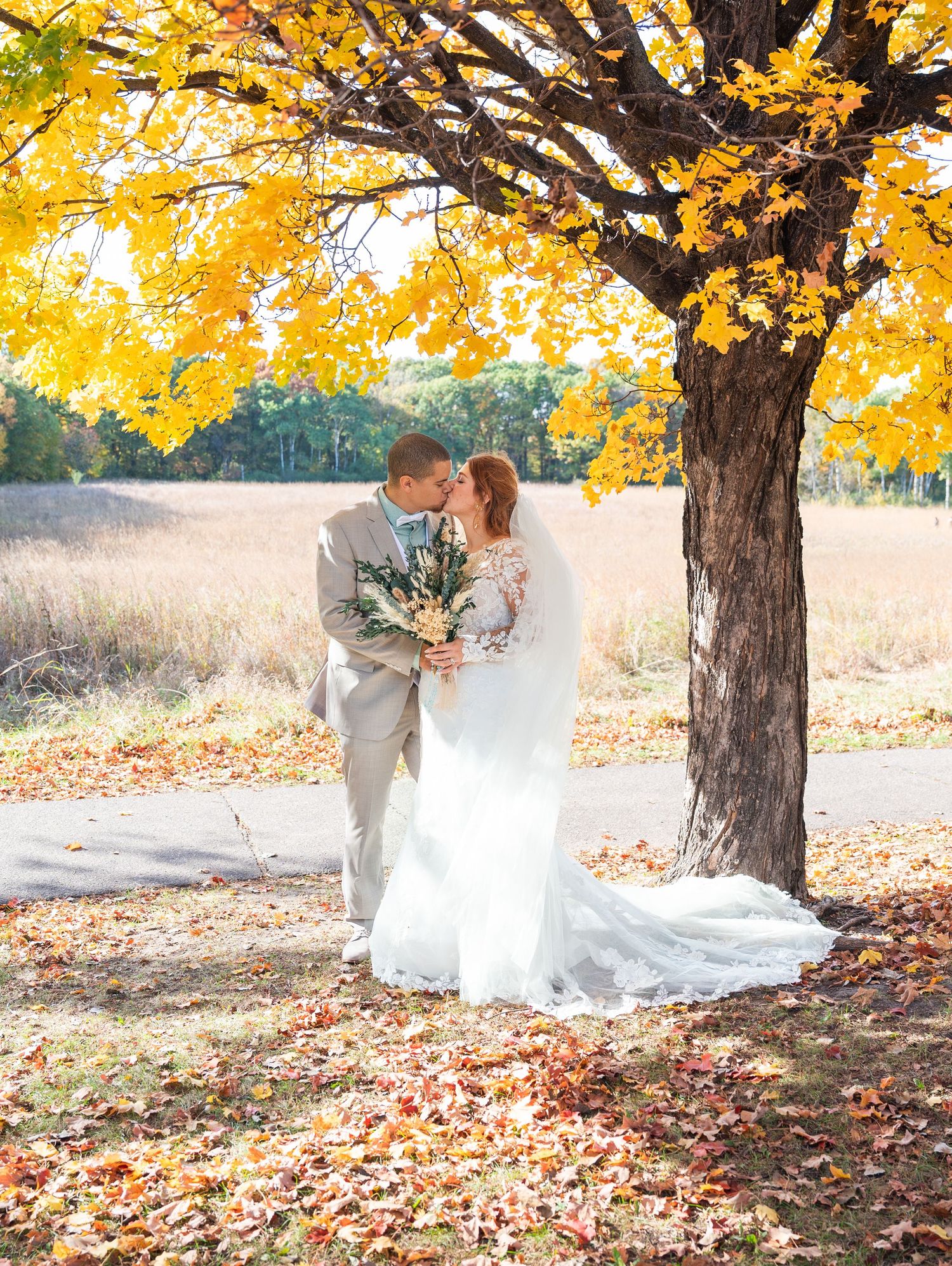 Bride and groom kissing under a golden autumn tree surrounded by fallen leaves in a romantic outdoor wedding photo.