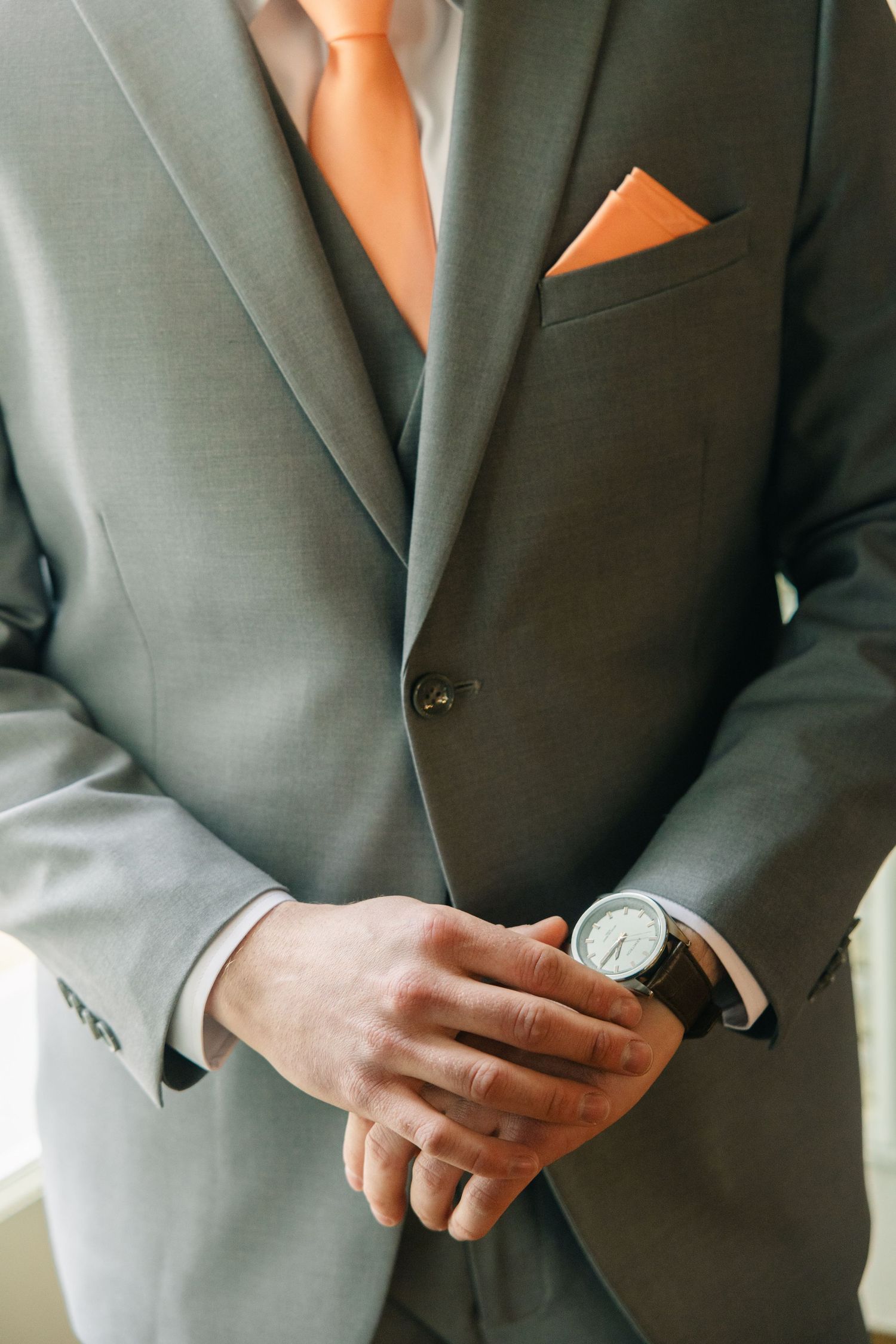 Man in gray suit with orange tie, matching pocket square, and brown leather watch, hands clasped.