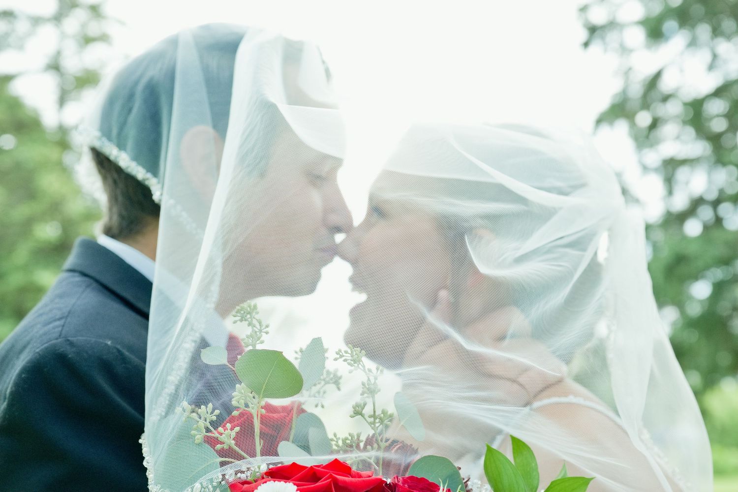 Newlyweds share a romantic kiss under a flowing veil with a red rose bouquet on their wedding day.