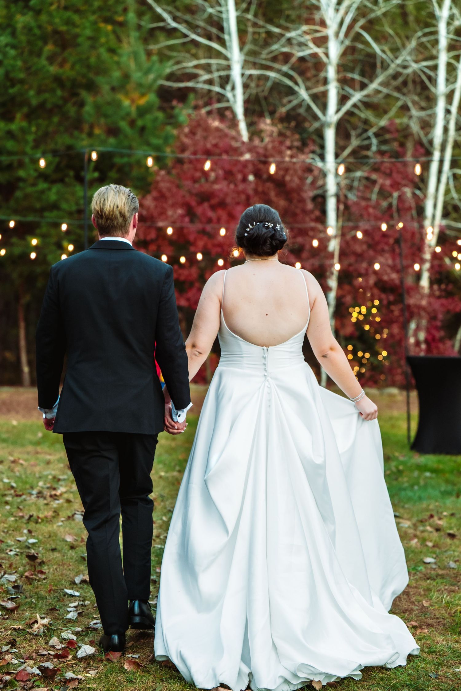 A couple walks through a garden setting with string lights and white birch trees.