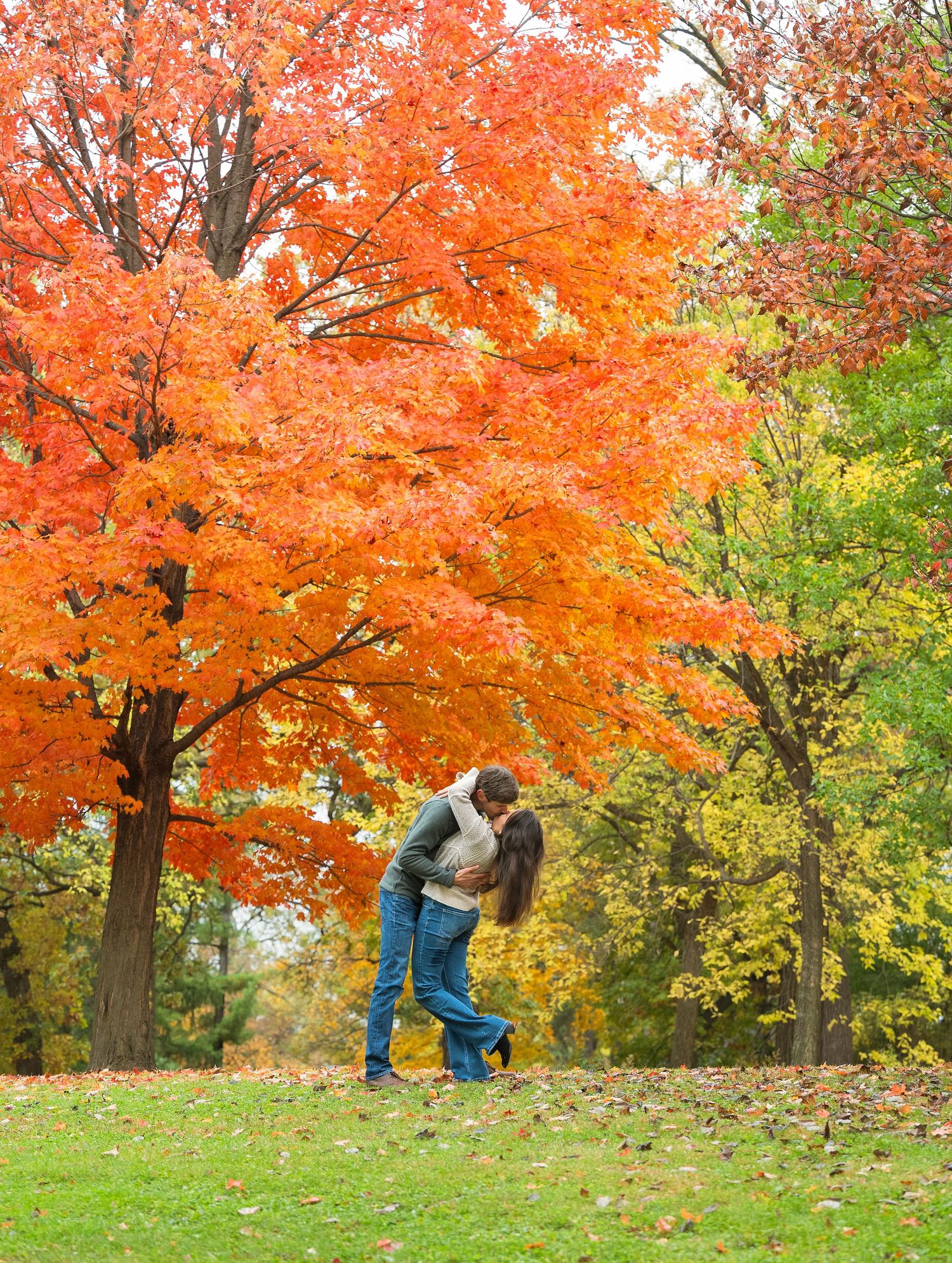 A couple dances and spins together beneath vibrant orange autumn maple trees in a park setting.