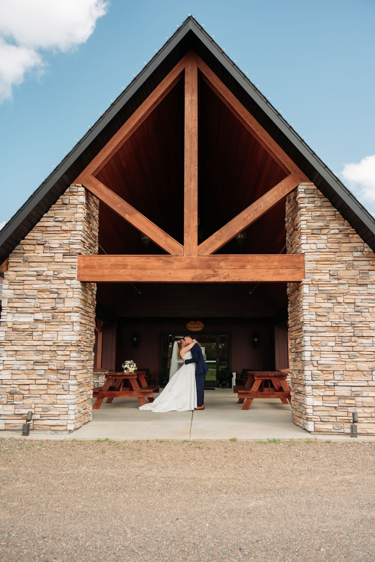 Stone pavilion with wooden beam accents and multiple covered sections under a bright blue sky. Pease Minnesota Wedding at Northern Lights Ba