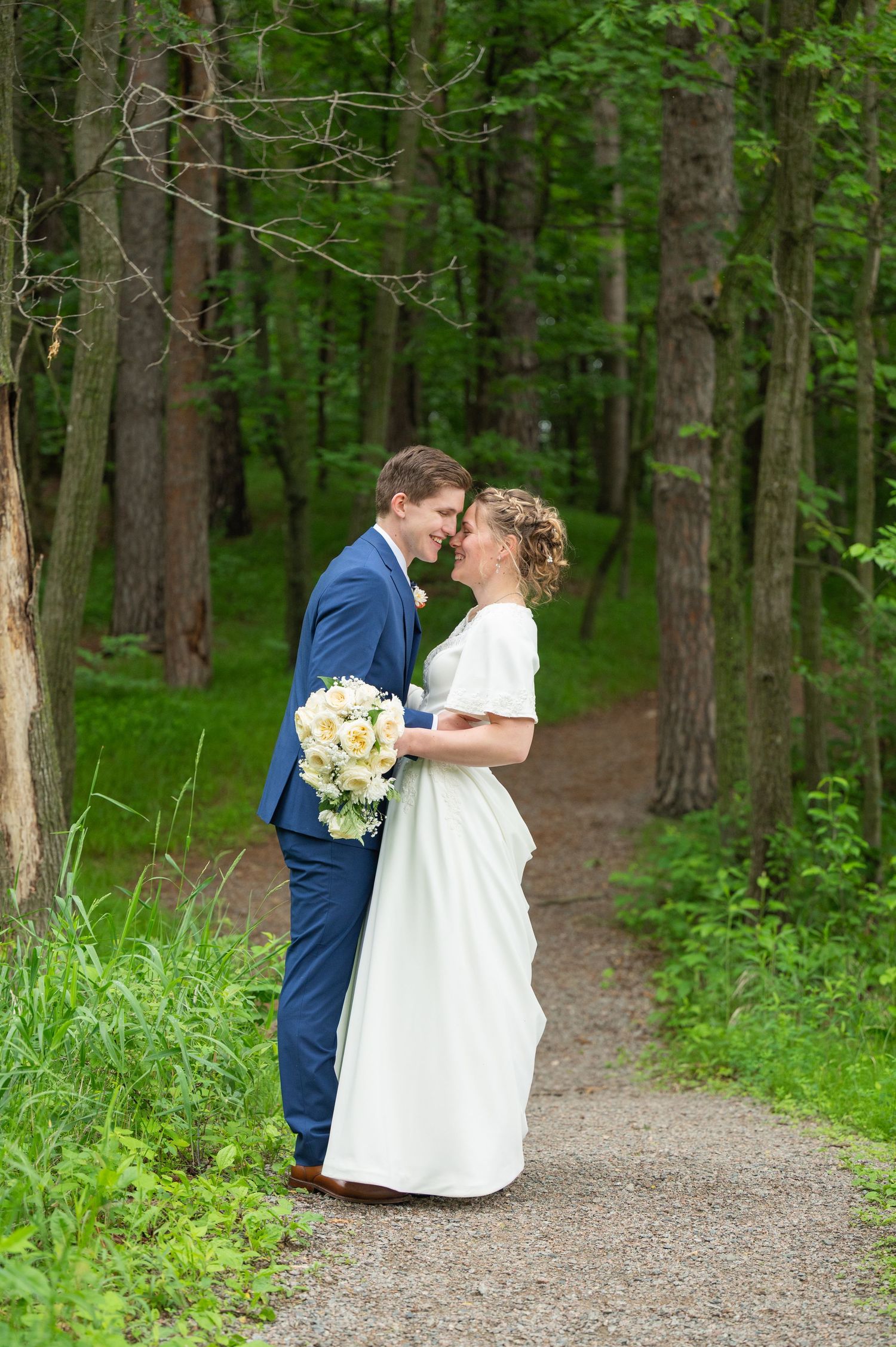 A wedding couple shares intimate moments on a wooded path surrounded by lush green forest.