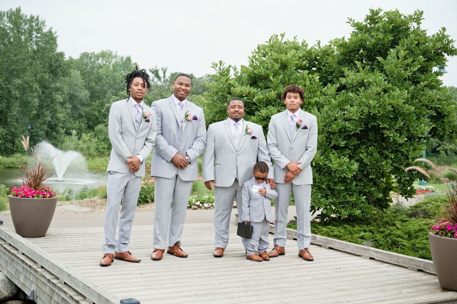 Groomsmen and ring bearer in light gray suits pose on wooden deck surrounded by lush greenery at outdoor wedding.