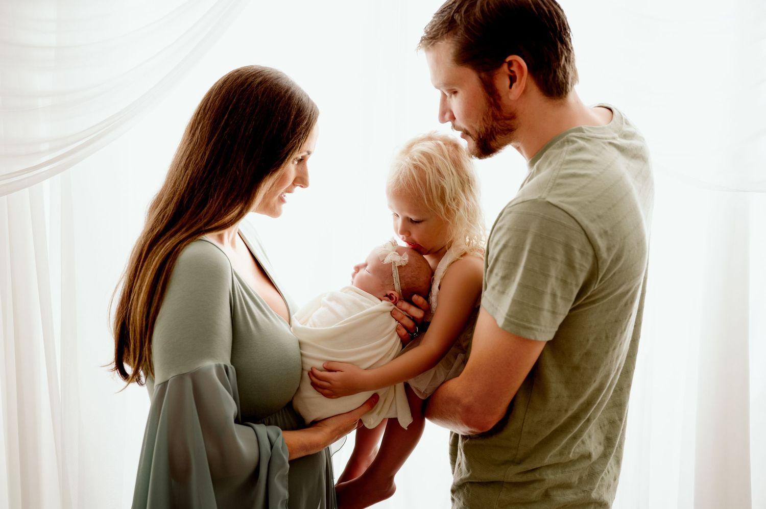 A family stands together near a white curtained window in soft natural lighting.