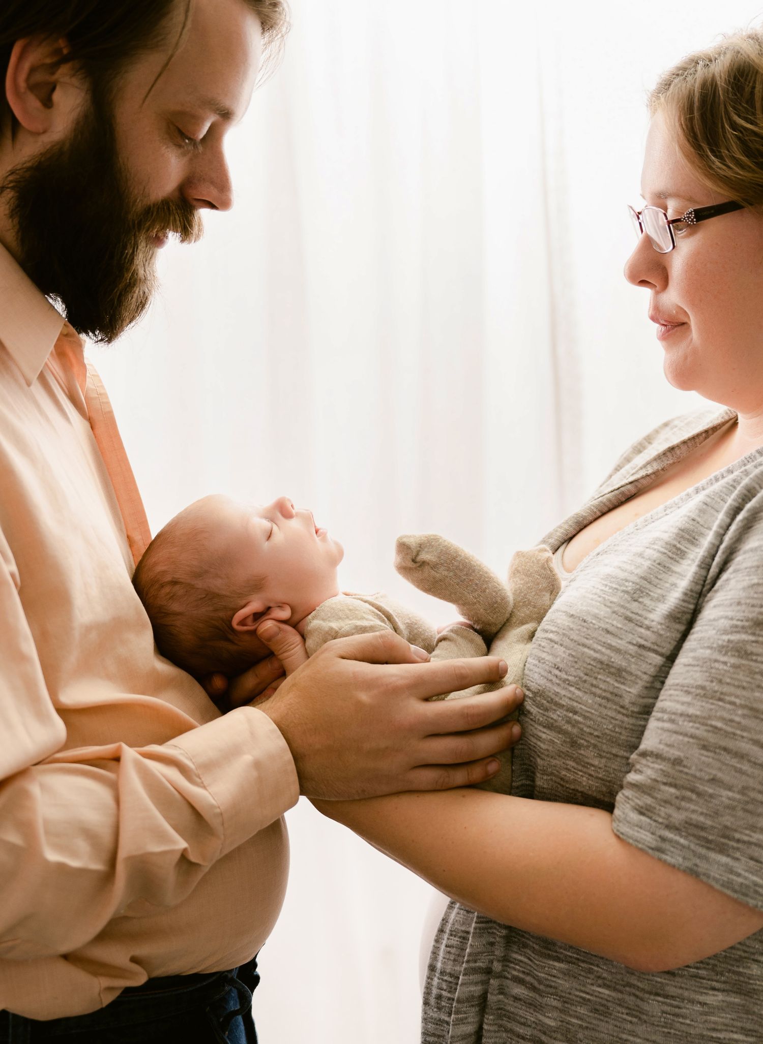 A couple holds their newborn baby in a softly lit room by a window.