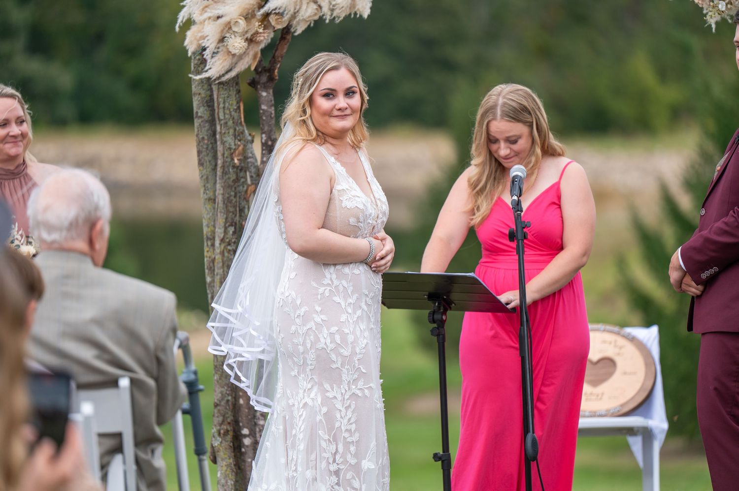 Wedding ceremony with bride in lace dress and person in bright pink dress standing at outdoor altar.