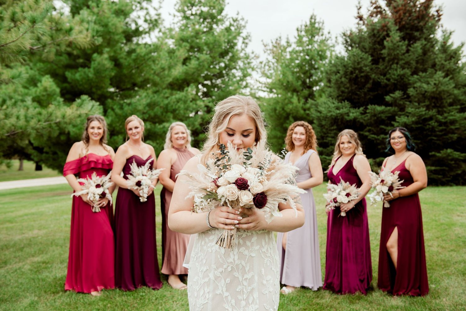 Bridal party in burgundy gowns stands outdoors with bouquets featuring deep red and white flowers.