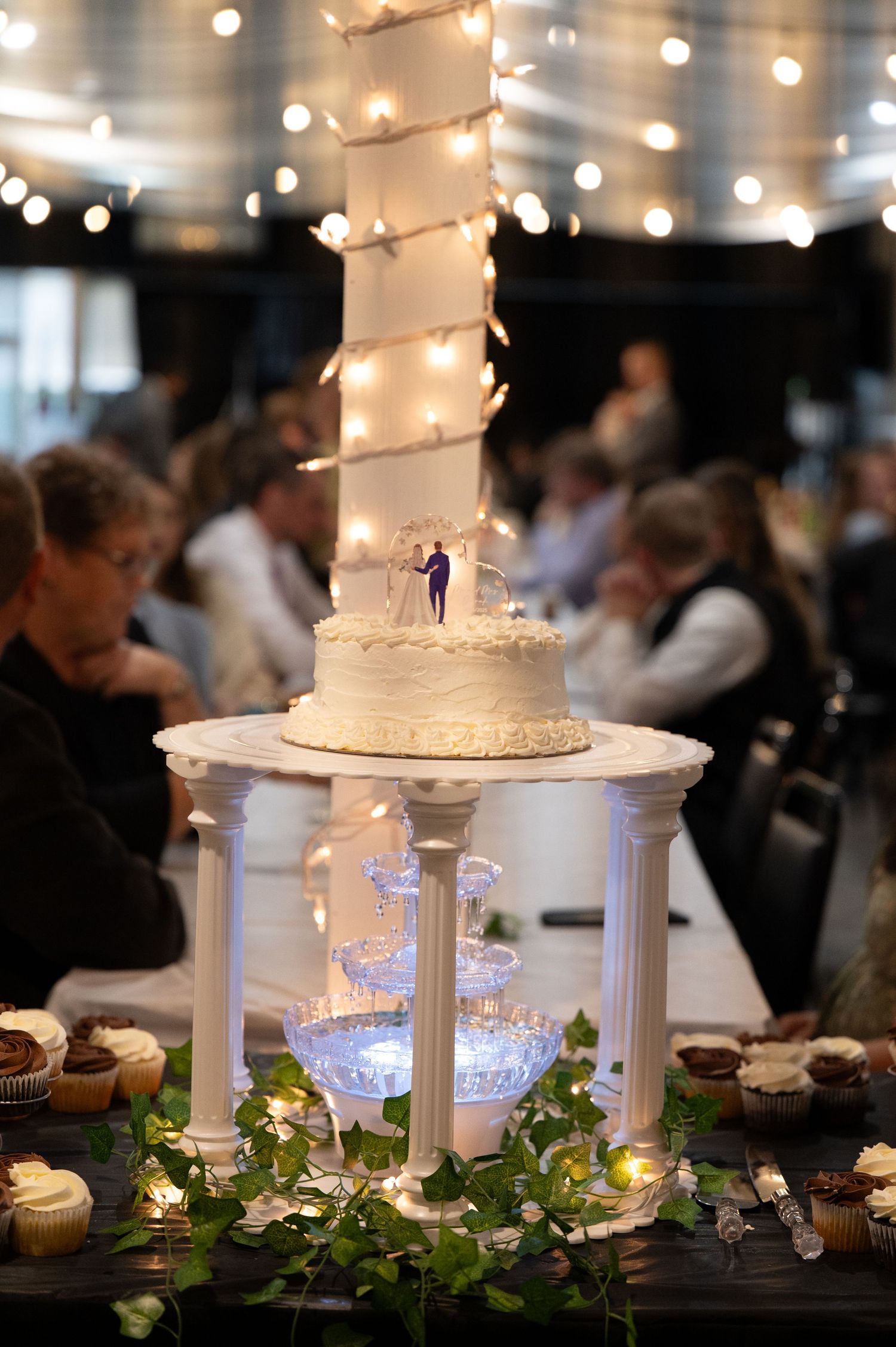 Elegant white wedding cake displayed on a lit pillar stand with string lights and greenery decorations at reception.