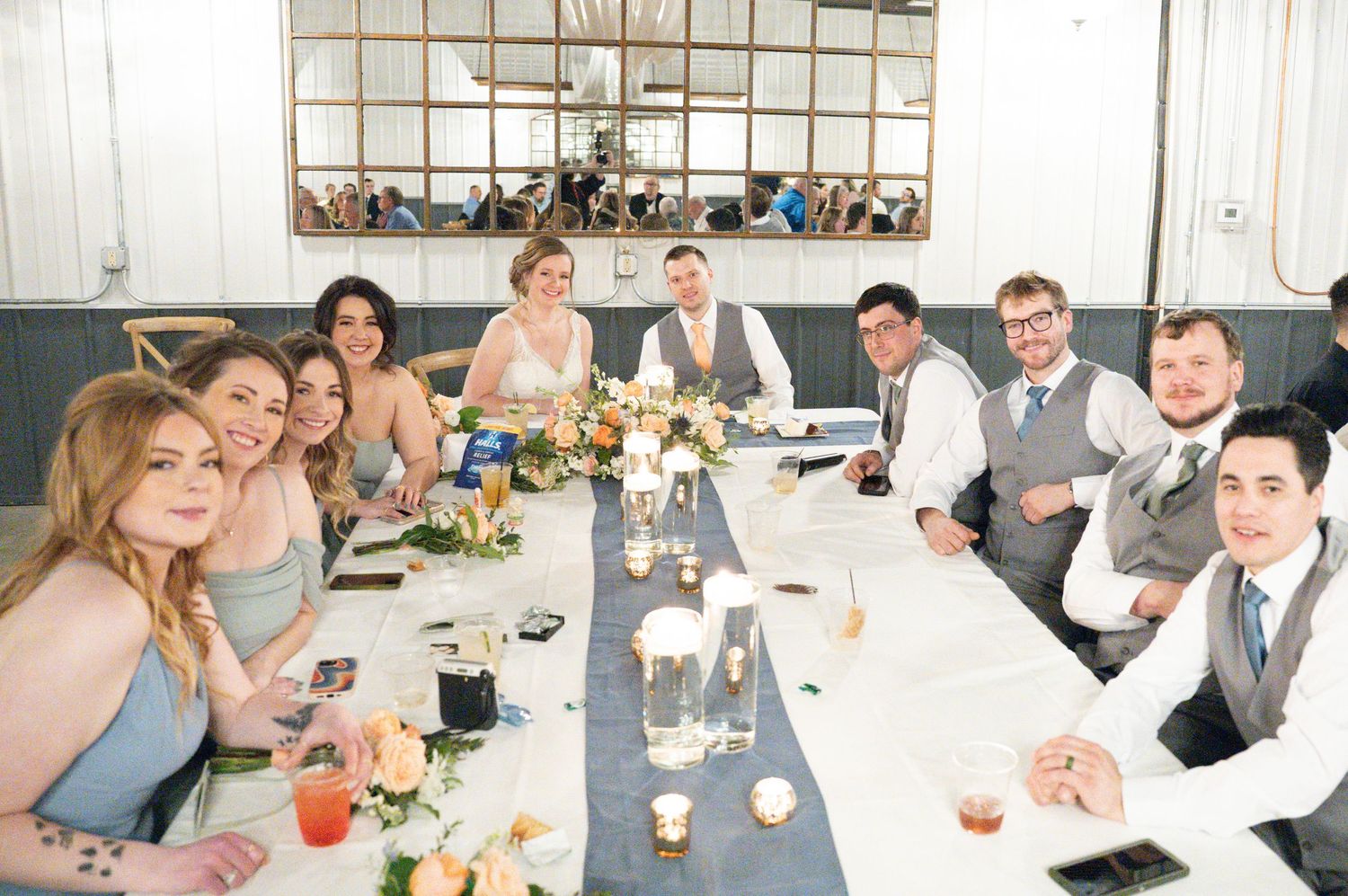 Wedding party smiles together at a long decorated reception table with candles and floral centerpieces.