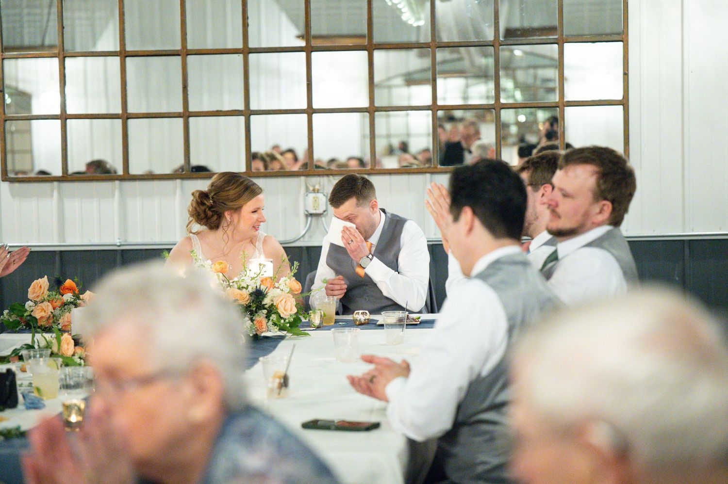 Bride and groom share emotional moment with wedding party at decorated head table during reception.